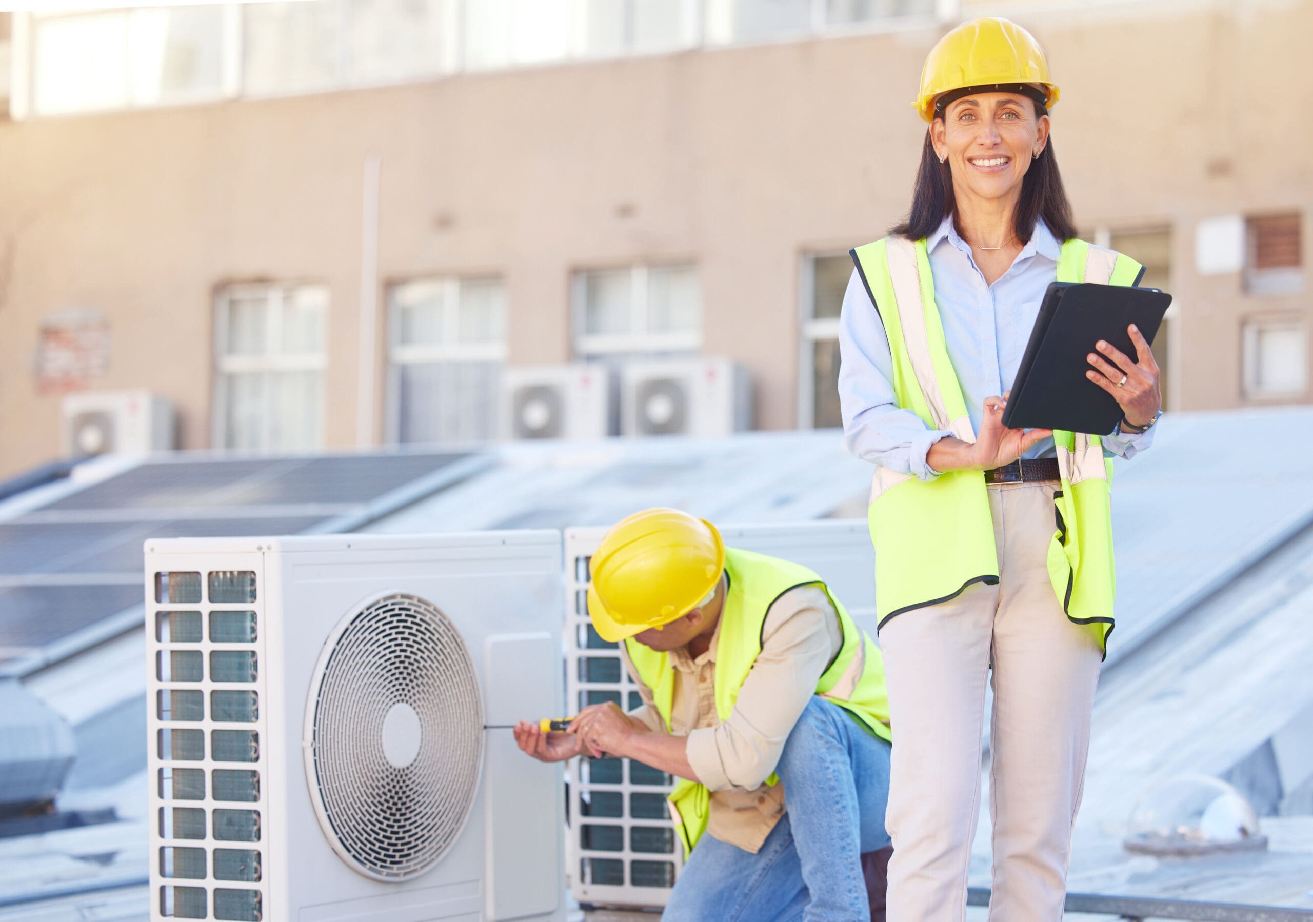 A woman is standing with a clipboard, facing towards while a man is bent down and working on the air system on a roof