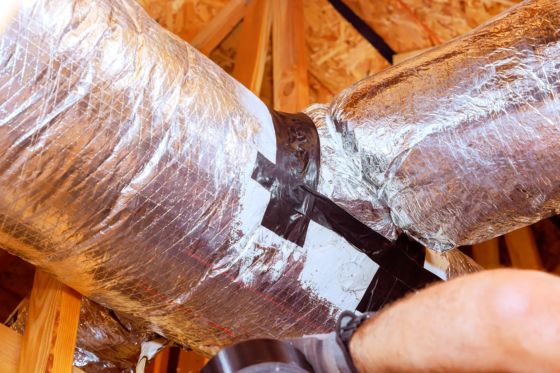 241036399_l Close-up of HVAC ductwork in an attic being sealed with foil insulation and black duct tape to prevent air leakage and improve energy efficiency.