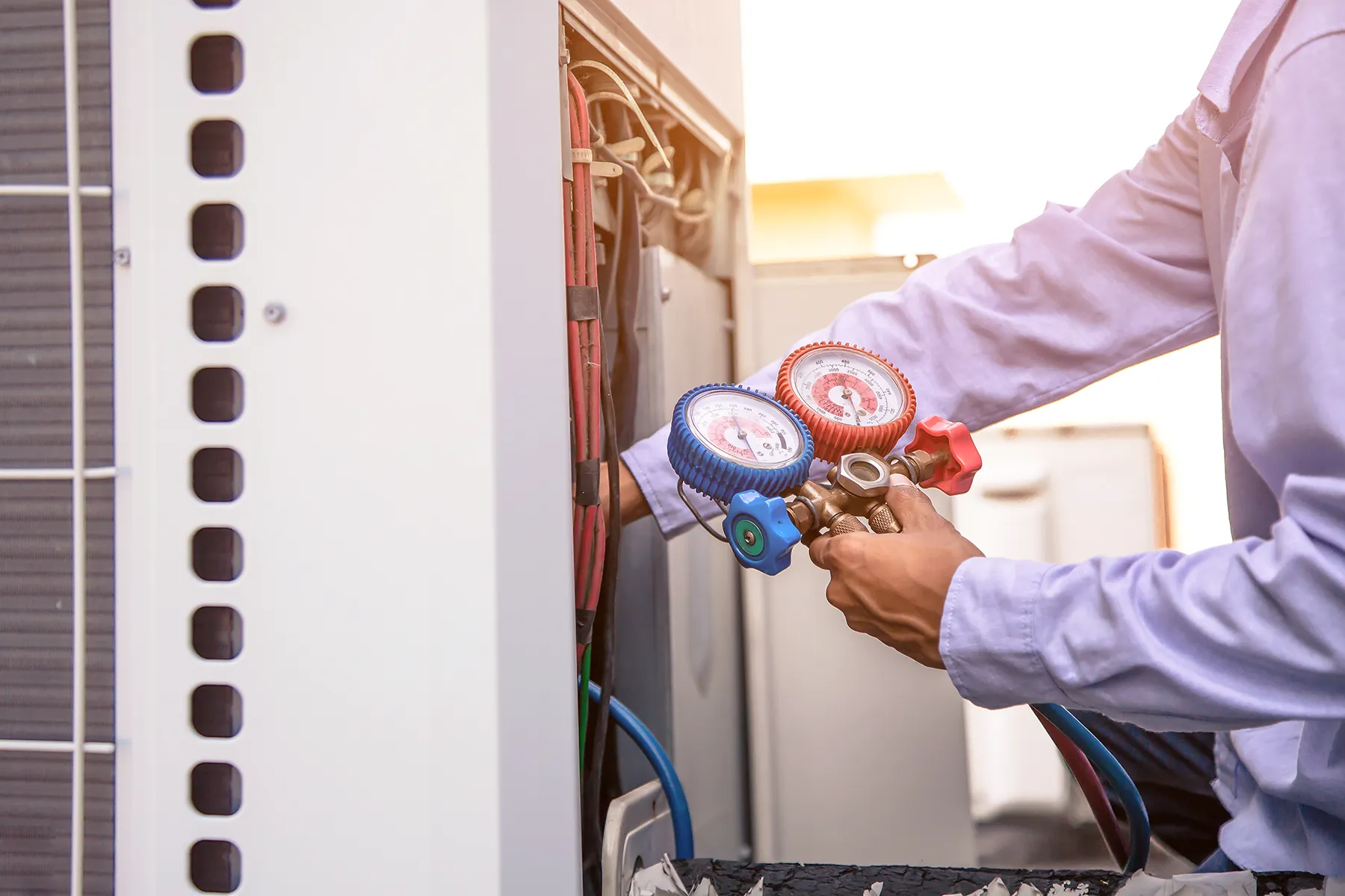 146455897_l HVAC technician using a manifold gauge to inspect and measure refrigerant pressure on an outdoor air conditioning unit during maintenance.