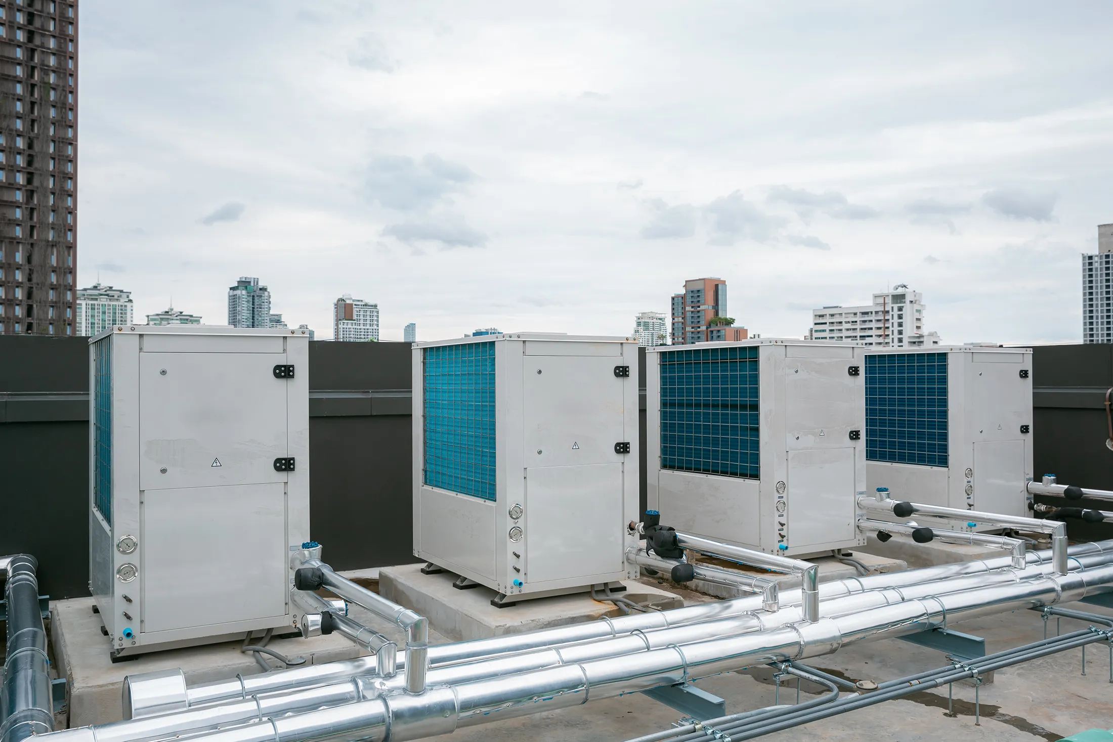air-conditioner-compressor-installed-in-old-buildi-2025-03-15-18-38-52-utc Commercial rooftop HVAC units with visible metal ductwork and city buildings in the background under an overcast sky.