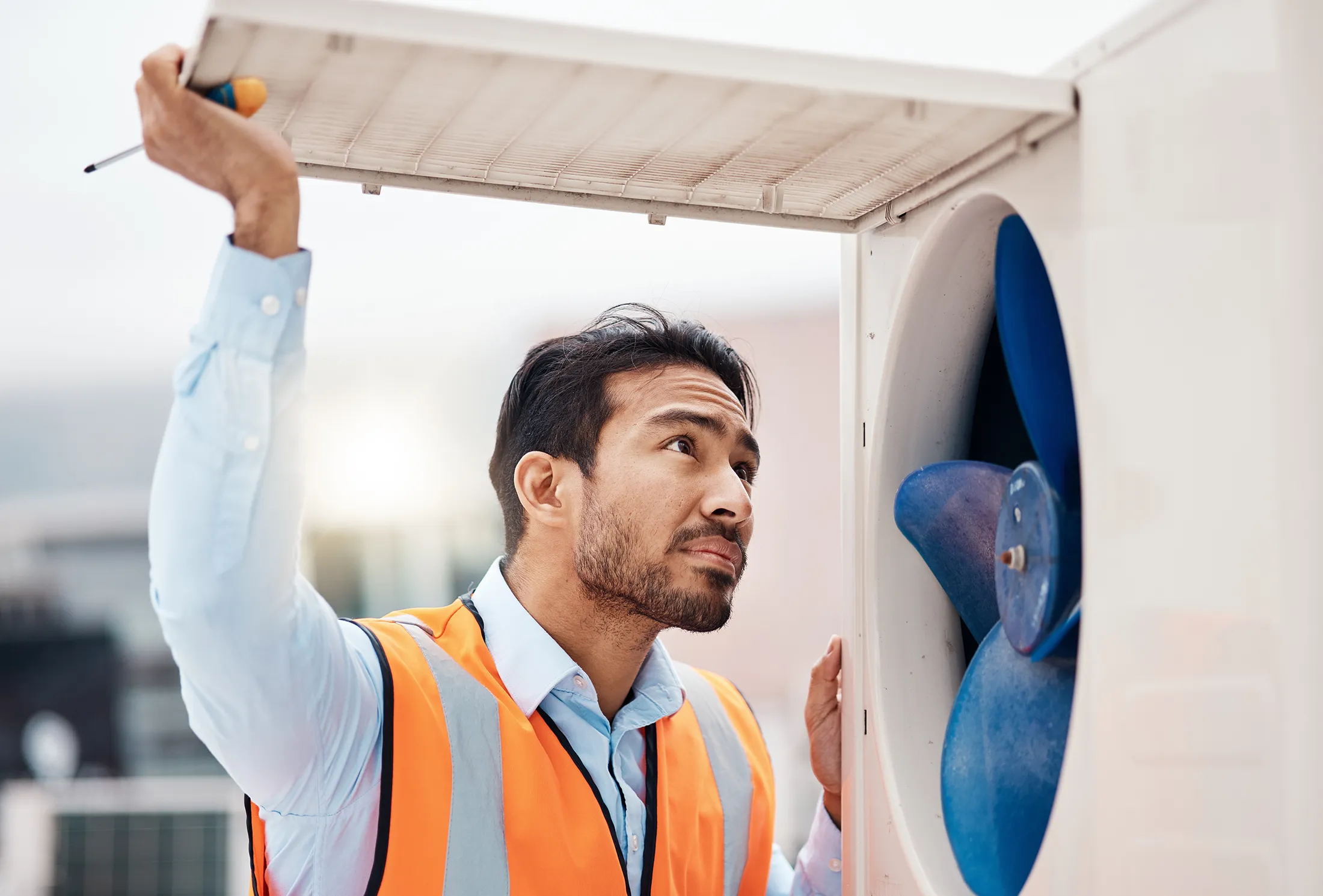 engineer-man-air-conditioning-and-maintenance-on-2025-04-06-11-47-46-utc HVAC technician inspecting a rooftop air conditioning unit with a blue fan while holding up the access panel.