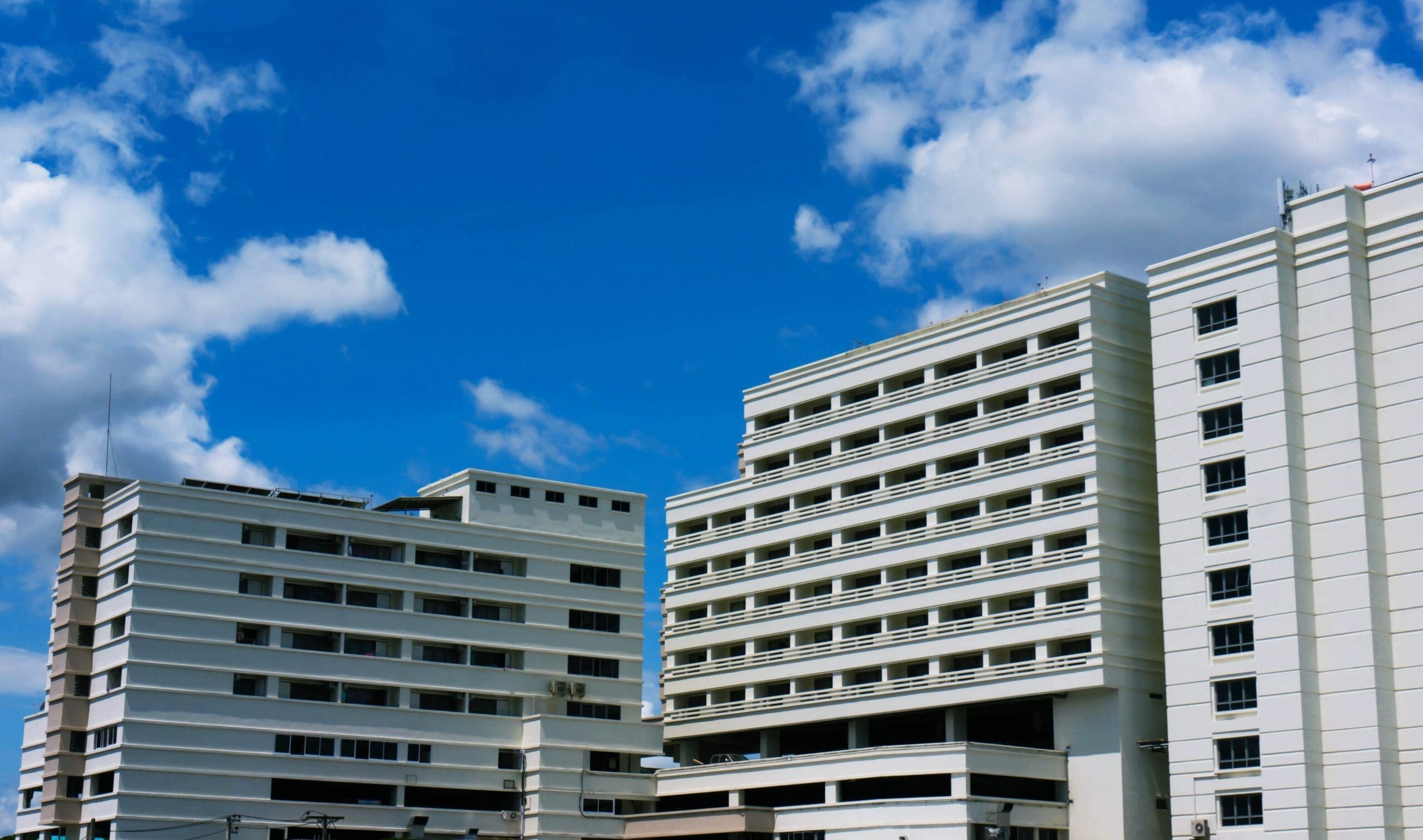Modern white apartment buildings beneath a bright blue sky with large white clouds during a sunny day.