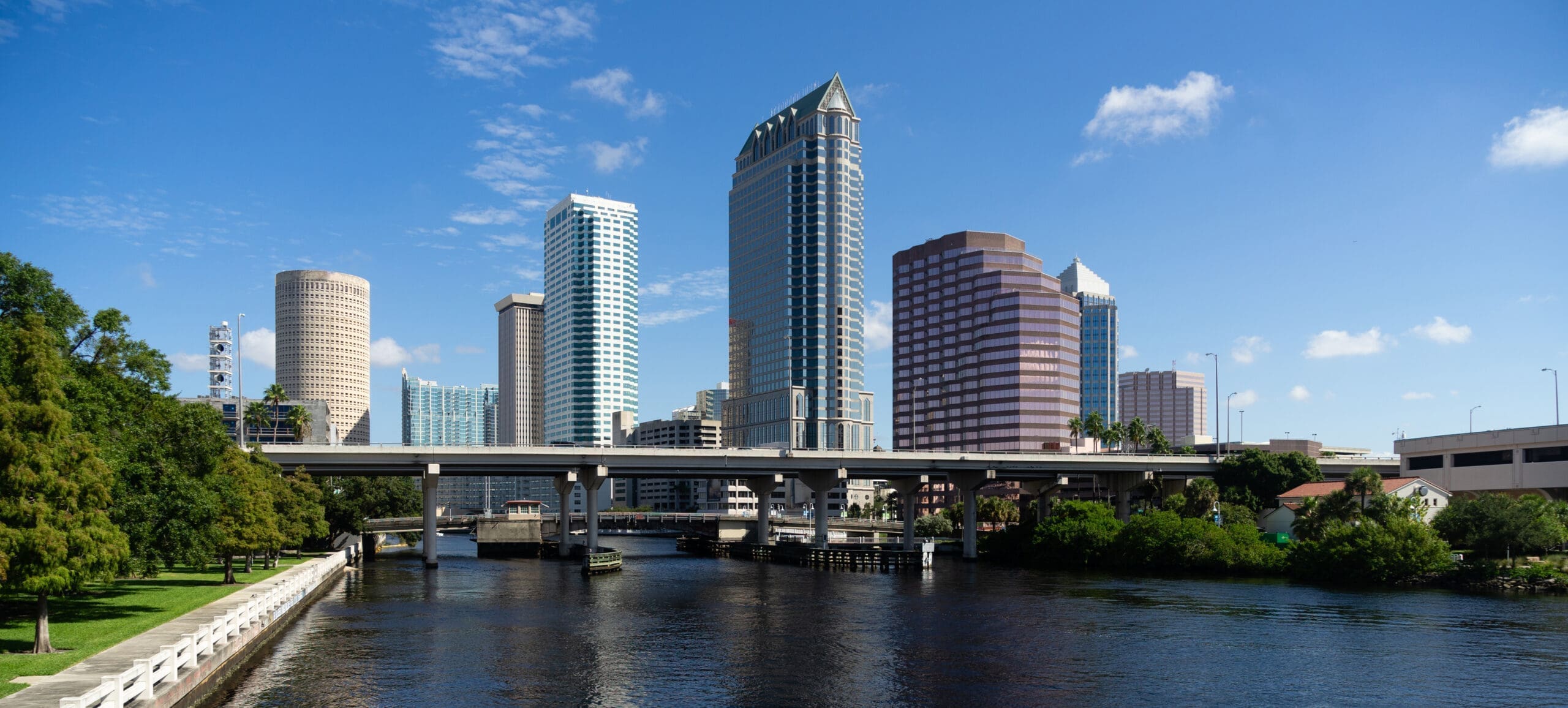 Downtown Tampa skyline featuring modern skyscrapers, a river canal, and elevated highway bridges under a clear blue sky.