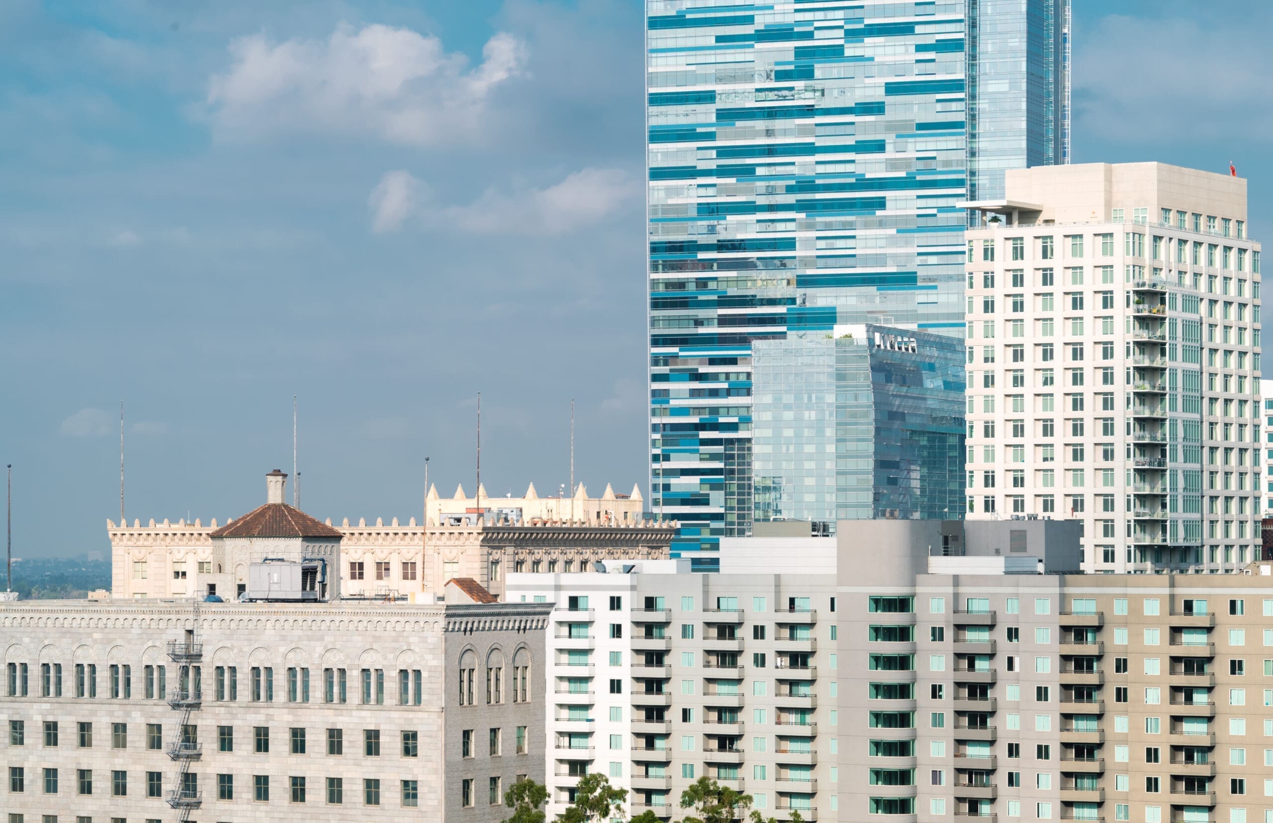 View of downtown Los Angeles buildings featuring a mix of historic architecture, modern apartment complexes, and reflective glass skyscrapers under a cloudy blue sky.