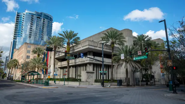 Urban city street featuring the Orlando Public Library building surrounded by palm trees, traffic lights, and nearby modern high-rise architecture under a blue sky.